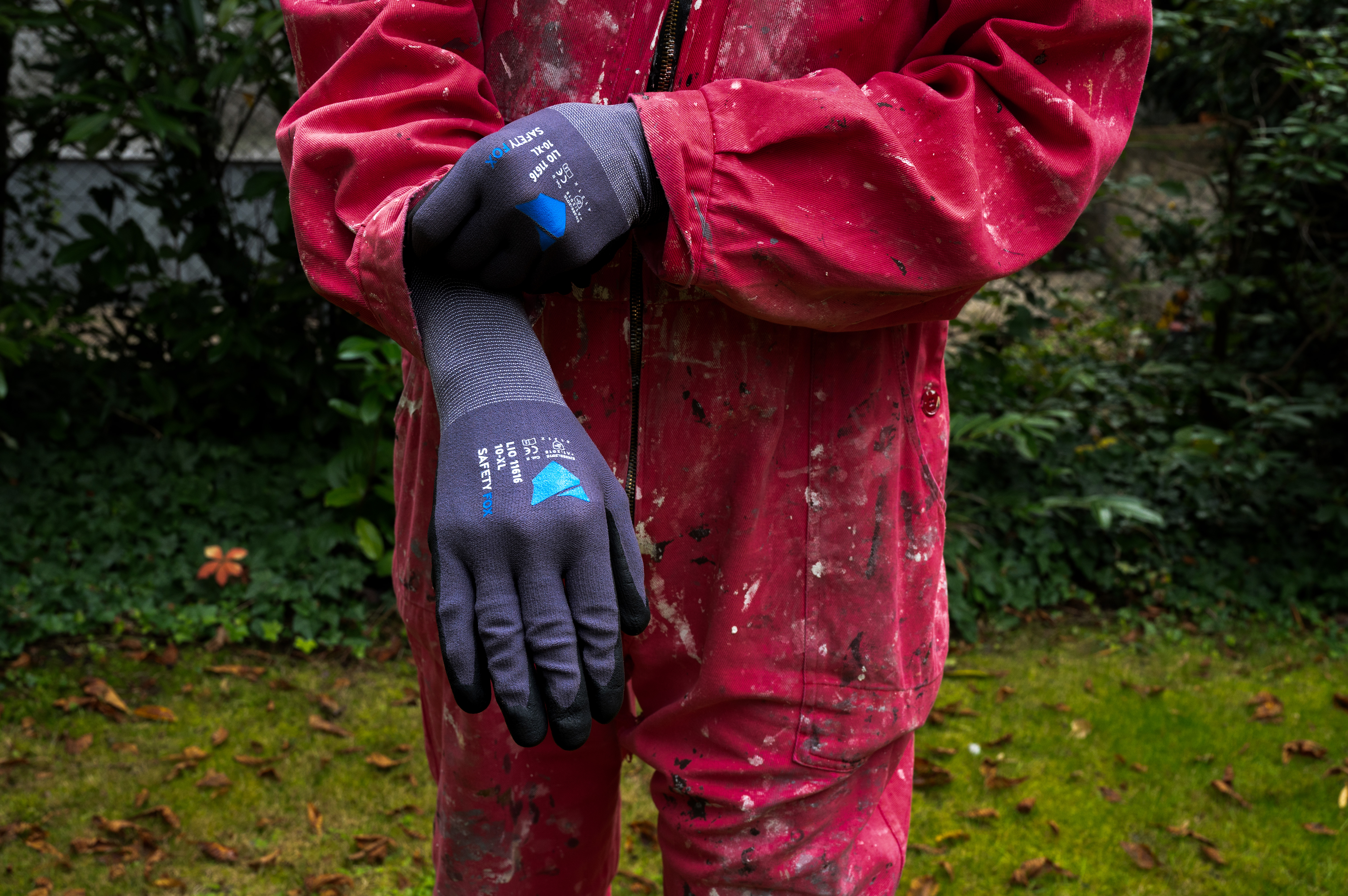 Person in verschmutztem roten Arbeitsoverall zieht graue Safety Fox Arbeitshandschuhe mit blauem Logo an